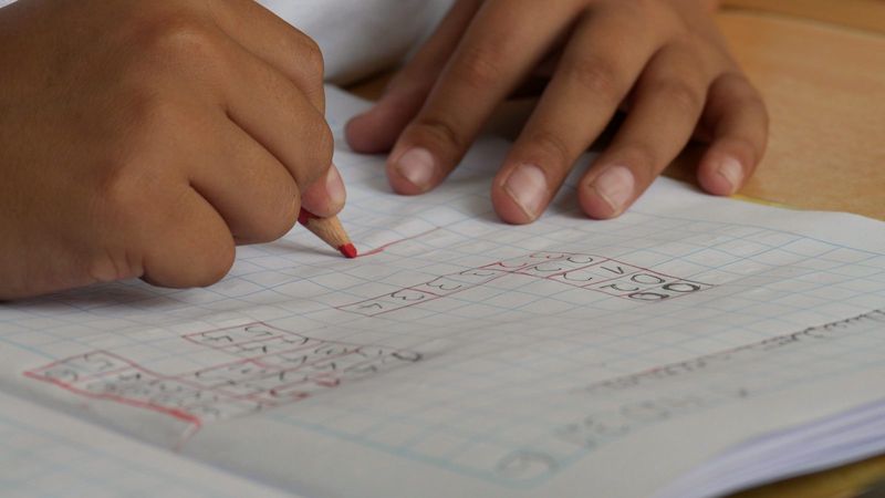 Close-up of a young student's hands working through sums on squared paper with a red pencil