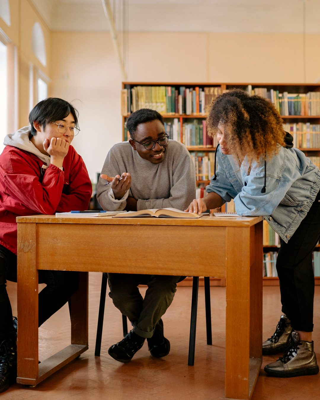 Three older students discussing an open book around a wooden table in a library